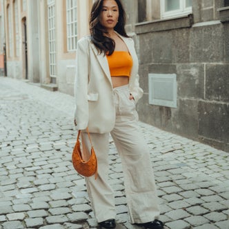 A woman wearing off white linen pants and blazer over an orange top against a background of a building and street