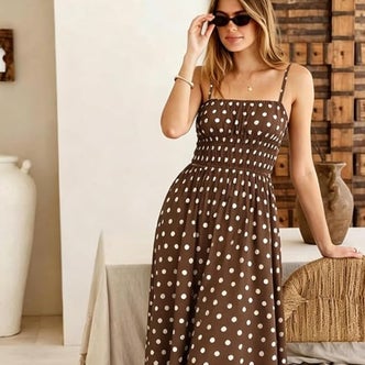 Woman wearing a brown polka dot smocked maxi dress with spaghetti straps, styled with black sunglasses and delicate gold jewelry in a warm, modern interior.