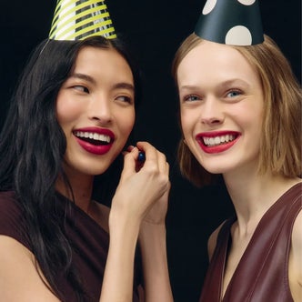 Two women wearing festive party hats smile at the camera with bold red lipstick, celebrating together against a black background