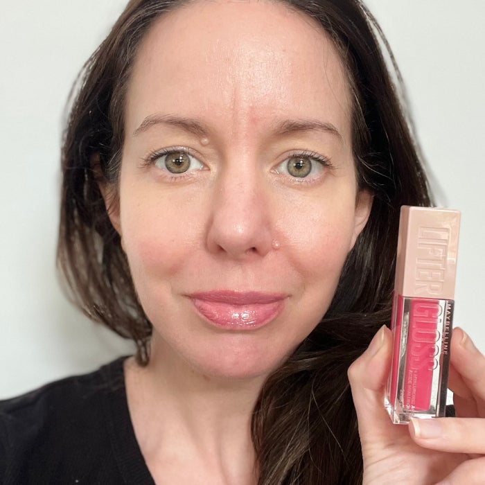 Brittany Brainard's face after applying Maybelline Lip Lifter Gloss and hand holding the lip gloss tube against a white background