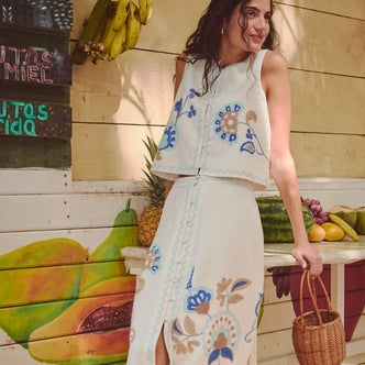 Woman wearing a white embroidered sleeveless top and matching midi skirt with blue floral detailing, holding a woven basket bag while posing beside a colorful tropical fruit stand