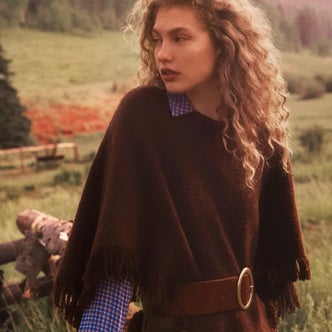 Anthropologie; Woman wearing a dark brown fringed poncho layered over a blue checkered shirt and cinched with a wide leather belt, posing in a sunlit countryside landscape.