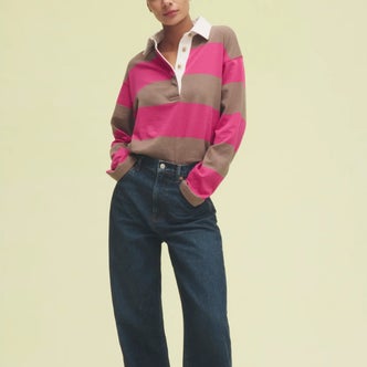 Model wearing a pink and taupe striped rugby shirt with a white collar, paired with dark-wash wide-leg jeans for a casual, preppy-inspired look against a soft yellow background