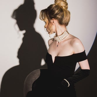 A woman wearing a black dress and pearl necklace looking at her shadow against a white background.