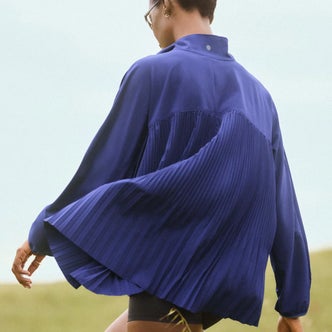 Person wearing a cobalt blue pleated performance jacket with flowing cape-like back panels, photographed from behind in an outdoor setting