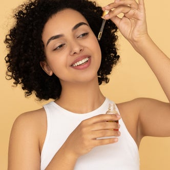 A woman holding a dropper of jojoba oil against a yellow background