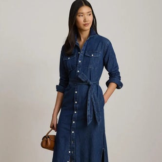 Woman wearing a dark-wash denim button-front midi dress with a tie waist and rolled sleeves, styled with a small brown handbag against a neutral studio background.