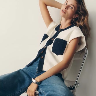 Woman wearing a cream sleeveless cardigan with contrasting navy trim and patch pockets, paired with relaxed blue jeans and silver jewelry, seated in a minimalist studio