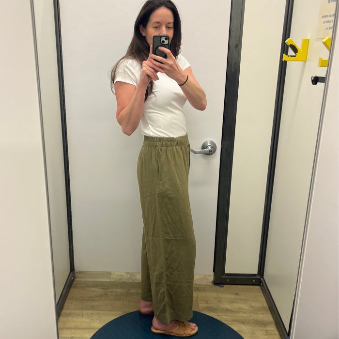 A side profile view of a woman in khaki linen pant and a white tee shirt against a white dressing room background