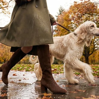 Person wearing an olive green trench coat and brown knee-high leather boots walking a cream-colored dog on a rainy autumn day, showcasing stylish waterproof footwear