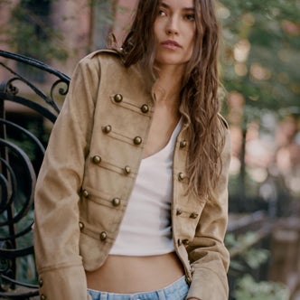 Woman wearing a tan military-style jacket with gold buttons over a white tank top