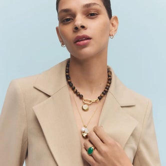 Model wearing a beige blazer styled with layered gold necklaces, a dark wood bead necklace, green statement ring, and small gold hoop earrings against a light blue background