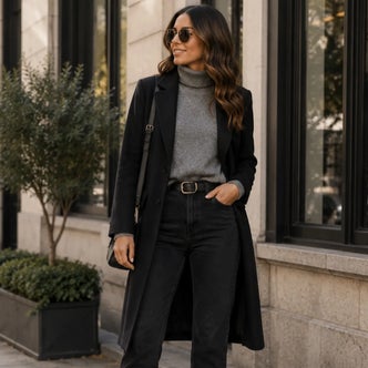 Woman wearing a black longline coat over a gray turtleneck sweater and black jeans, accessorized with sunglasses and a shoulder bag.