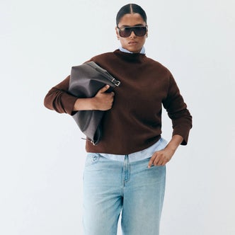 Woman wearing a chocolate brown sweater layered over a blue striped shirt with light-wash jeans, oversized sunglasses, and a dark leather clutch.