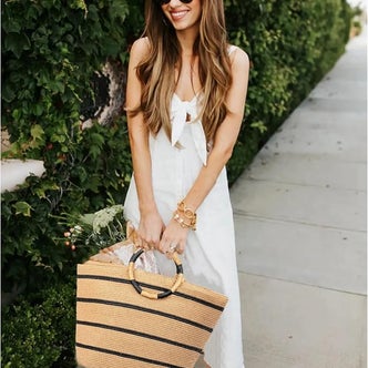 Woman wearing a white tie-front maxi dress and black sunglasses, carrying a striped woven straw tote bag while walking along a tree-lined sidewalk.