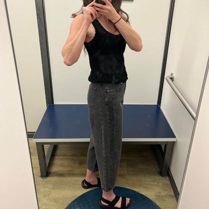 A side profile view of Brittany Brainard wearing gray Old Navy High Waist Ankle Barrel Jeans with a black tank top in a white dressing room.